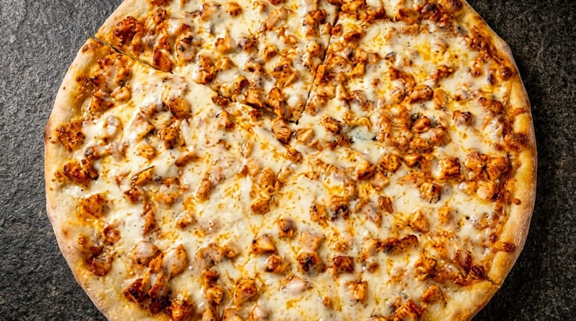 A whole chicken pizza, topped with melted cheese and diced chicken, on a dark countertop.